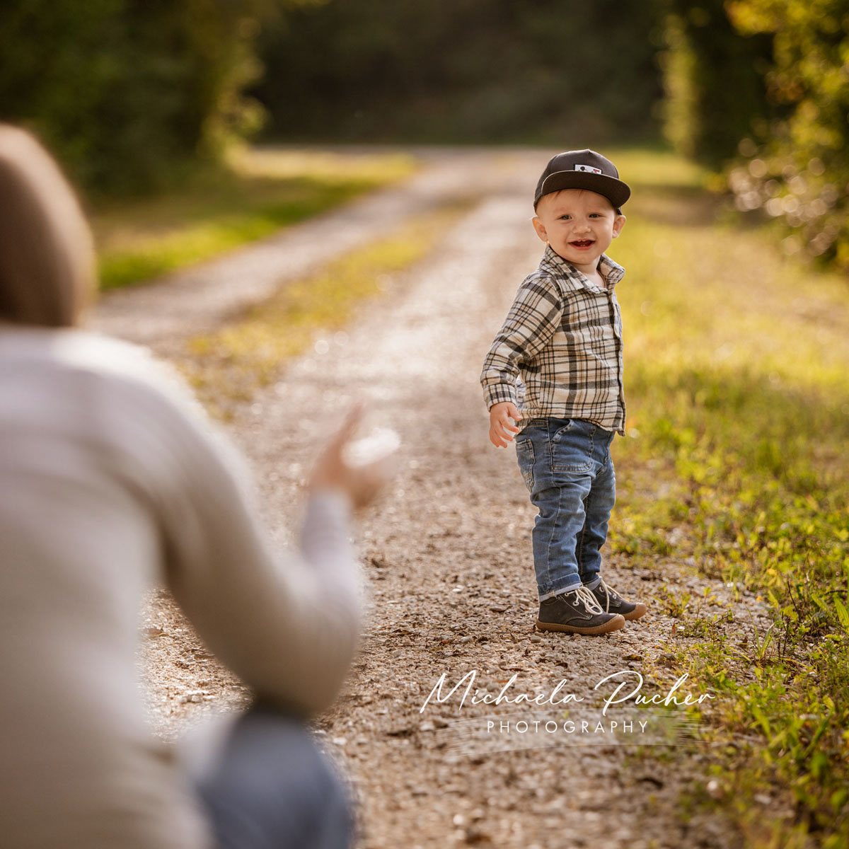 Lachender kleiner Junge mit Kappe steht auf einem sonnigen Feldweg und blickt zu einer erwachsenen Person im Vordergrund.